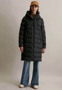 Svart puffjakke med hette, glidelås foran og snøring. Har en lang, quiltet design og er parret med vide blå jeans.