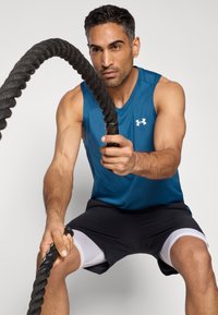 Maglietta atletica senza maniche blu e pantaloncini neri con dettagli bianchi. La persona impugna una fitta corda da battaglia nera, concentrata sull'attività.