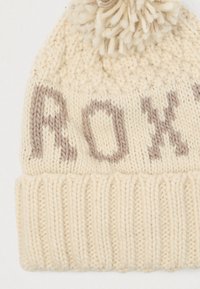 Pletená čiapka v odtieňoch off-white s svetlohnedým nápisom "ROXY." Má zaoblenú korunku a pom-pom na vrchu; ribbovaný lem na spodnej časti.