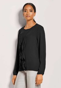 Zwarte blouse met lange mouwen en verticale plooien aan de voorkant, gemaakt van een zachte stof. Lichtgewicht met een ronde halslijn, gecombineerd met lichtgekleurde broek.