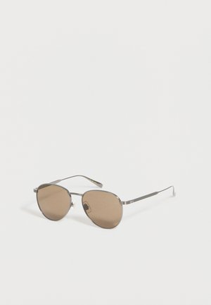 Lunettes de soleil avec monture métallique argentée et verres ovales teintés marron reposant sur une surface blanche.