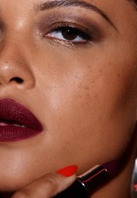 Rossetto opaco color bordeaux scuro applicato sulle labbra, accompagnato da un ombretto marrone lucido e smalto rosso brillante su pelle liscia.