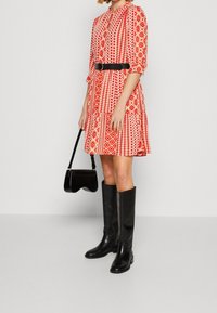 Robe à motifs rouges et blancs avec des manches trois-quarts et une ceinture noire. Portée avec des bottes noires jusqu'aux genoux et tenant un petit sac à main noir.