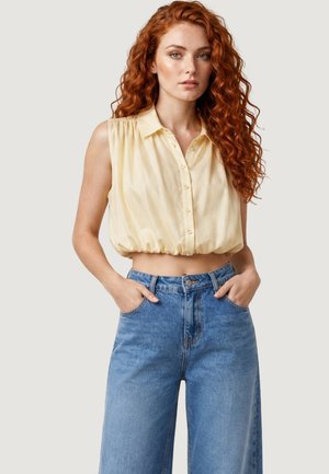 Femme aux longs cheveux roux bouclés portant un crop top jaune sans manches boutonné et un jean bleu taille haute à jambes larges, mains dans les poches.