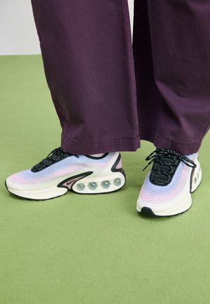 Voeten met witte sneakers en een pastelachtige gradiënt, voorzien van zichtbare luchtkussens, gecombineerd met losse donkerpaarse broeken op een groene vloer.