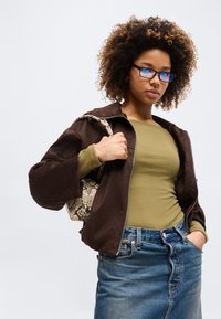 Jeune femme aux cheveux bouclés et lunettes, portant une veste marron, un haut vert, une jupe en jean, tenant un sac à main à motifs sur l'épaule.