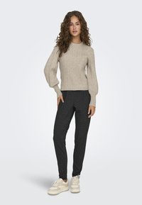Pull en tricot beige avec un motif texturé, manches légèrement bouffantes, associé à un pantalon noir grisâtre ajusté. Le mannequin porte des baskets crème.