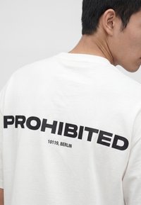 Mężczyzna w białej koszulce z czarnym napisem "PROHIBITED" i mniejszym tekstem "10119, BERLIN" na plecach, patrzy w bok.