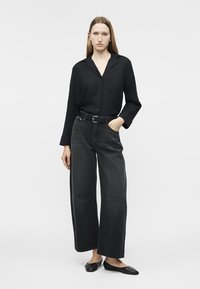 Kvinne står med én hånd i lomma, iført en svart skjorte med knapper, svarte vide jeans, svart belte og svarte flate sko.