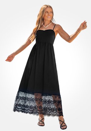 Vrouw in een mouwloze zwarte maxi-jurk met kanten zoom, sandalen en gelaagde kettingen, poseert met uitgestrekte armen en glimlacht.