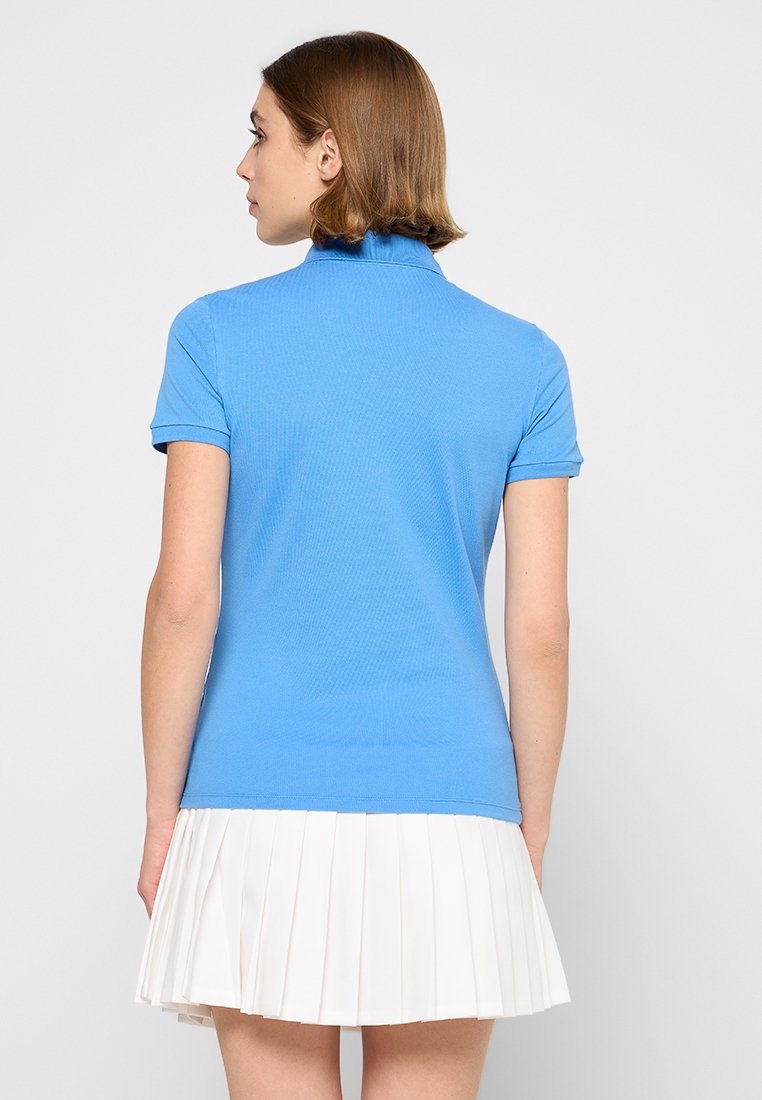 Femme aux cheveux bruns courts de dos, portant un polo bleu à manches courtes et une jupe plissée blanche.