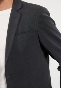 Herenblazer van zwart katoen met een gestructureerde stof, enkele borstzak en subtiele details. Witte t-shirt zichtbaar onder de jas.