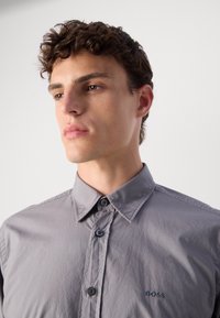 Camicia grigia con bottoni, realizzata in tessuto liscio. Presenta un colletto classico, due bottoni sul davanti e un piccolo logo vicino all'orlo.
