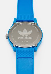 Hodinky s modrým silikónovým remienkom a nerezovým zadným krytom s gravírovaným logom a detailmi. Textúrovaný remienok a strieborné kovové prvky.