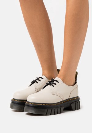 Chaussures à lacets beige clair avec une tige en cuir lisse, une semelle plateforme en caoutchouc noir avec des détails rainurés et des accents de couture jaunes.
