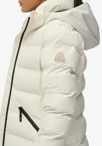 Mujer vistiendo una chaqueta de plumas blanca con capucha y un bolsillo con cremallera negra en ángulo, mostrando un parche con el logo de Superdry en la manga.