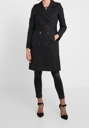 Woman wearing a black belted trench coat, black skinny jeans, fishnet socks, and black high heels with ankle straps and bows.