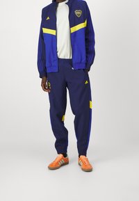 Adidas tepláková souprava v námořnické modři s žlutými a modrými akcenty. Obsahuje bundu se zipem a kalhoty s elastickým pasem. Doplněno o oranžové a černé tenisky.