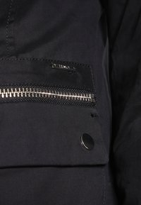 Detalj av svart jacka med en dragkedjeficka, silverfärgad dragkedja, synlig tygtextur och en rund svart tryckknapp. Minimal varumärkesexponering.