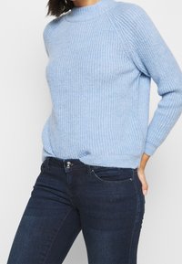 Lichtblau trui van gebreid materiaal met een hoge hals, geribbelde textuur en lange mouwen, gecombineerd met donkerblauwe jeans.