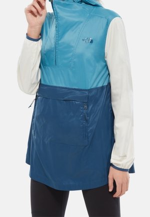 Vrouw draagt een blauw two-tone mouwloze jas over een witte top met lange mouwen, houdt de rits van de jas bij de kraag vast.