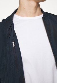 Námořnická bomber bunda s kapesním zipem, s žebrovaným límcem a hladkou texturou, nošená přes jednoduché bílé tričko.