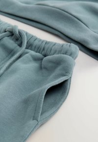Miękkie sweaty w kolorze mięty z elastycznym paskiem w talii, sznurkiem do regulacji i boczną kieszenią, leżące płasko na białej powierzchni z częściowo widoczną pasującą bluzą.