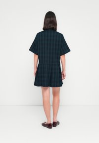 Kurzärmeliges Kleid aus dunkelgrünem Karostoff, ausgestellt geschnitten, mit einem plissierten Rock und geradem Rückendesign. Kombiniert mit dunkelroten Schuhen.