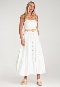 Biała sukienka maxi z ryftową górą, warstwowym krojem, guzikami z przodu i plecionym, brązowym paskiem. Noszona z kremowymi sandałami na platformie.