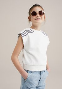 Fehér, rövid ujjú pulóver, tengerészkék hullámos hímzéssel a vállakon, világoskék csíkozott farmerral párosítva. A modellen kerek napszemüveg van.
