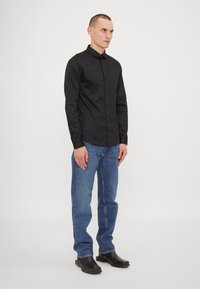 Camisa negra de manga larga con botones, diseño ajustado, textura suave, combinada con jeans rectos de tiro medio en azul claro y zapatos negros sin cordones.