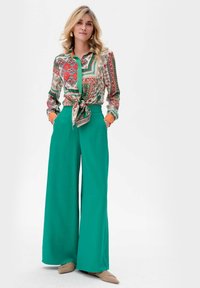Kleurrijke blouse met patroon en een knoop in de taille, gecombineerd met hoge, wijde groene broek en beige puntige flats.