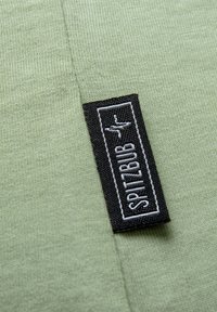 Light green textile with a smooth texture, featuring a black fabric tag with white stitching that reads "SPITZBUB."