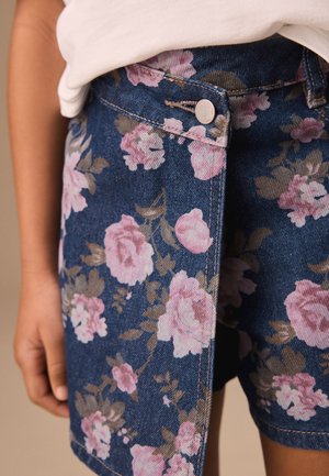 Uzorak plavih traper kratkih hlača s ružičastim i smeđim cvjetnim uzorkom nošen s bijelim topom, prikazujući gumb i ruku koja počiva sa strane.