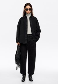 Zwarte denim jacket met studs, grijzige coltrui, bijpassende zwarte wijd uitlopende broek en elegante zwarte enkellaarsjes. Houdt een zwarte tas met franjes vast.