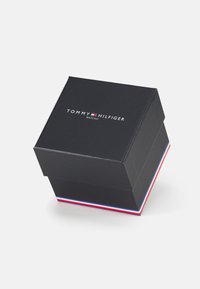 Black square box with a lid, displaying the "TOMMY HILFIGER" logo. Features a red, white, and blue striped accent at the base.