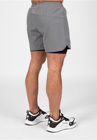 Grijze sportshorts met een zwarte binnenvoering, elastische tailleband en zijzakken, gecombineerd met witte en zwarte sneakers. De textuur lijkt glad.