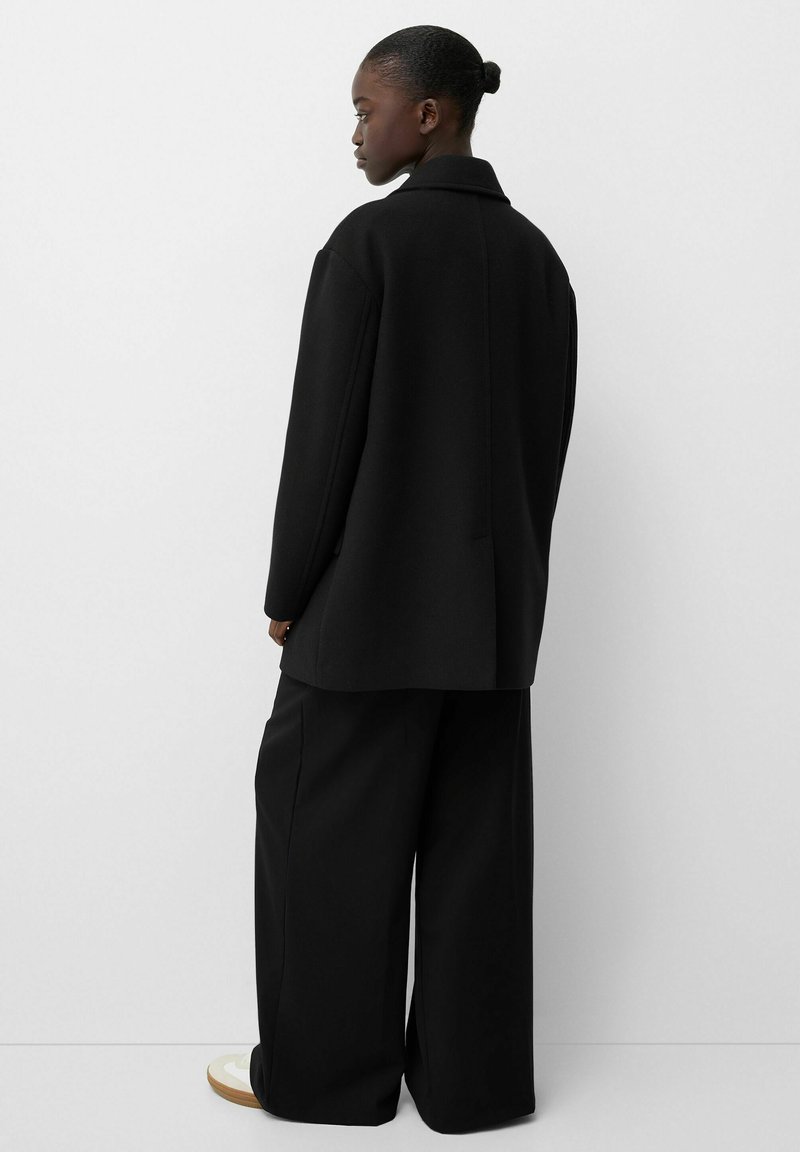 Schwarzer, übergroßer Mantel mit Reverskragen, aus einem strukturierten Stoff gefertigt. Verfügt über eine lockere Silhouette und weite schwarze Hosen.