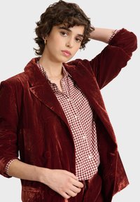 Czerwony aksamitny żakiet z dwoma guzikami, noszony na burgundowej bluzce w białą krateczkę. Kręcone włosy, ręka na głowie.