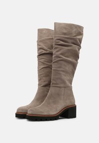 Botas de ante taupe hasta la rodilla con tacones de bloque, suelas negras con estrías y cañas suaves y holgadas sobre un fondo blanco.