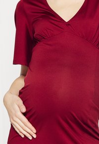 Personne enceinte portant une robe rouge tenant délicatement son ventre d'une main contre un fond clair uni.