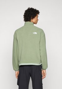 Roheline fleece-svetar kõrge kraega, ribikurdega mansetid ja lõdva lõikega. Seljal on valge logo ja kontrastne serv.