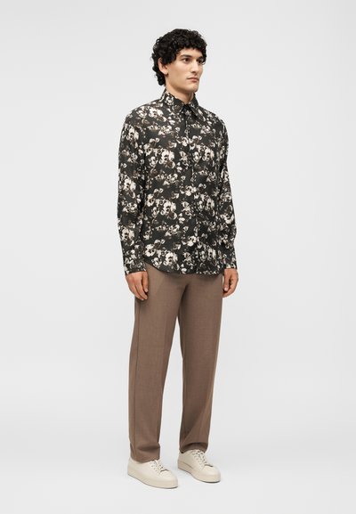 Άνδρας στέκεται φορώντας μαύρο πουκάμισο με κουμπιά και λουλούδια, καφέ παντελόνι και λευκά αθλητικά παπούτσια μπροστά σε απλό ανοιχτό γκρι φόντο.