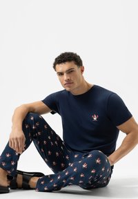 Man die op de grond zit, gekleed in een marineblauwe t-shirt met een cartoonhaan, en een bijpassende broek met patroon en zwarte sandalen.