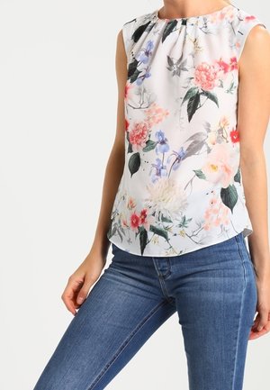 Haut blanc sans manches avec imprimé floral multicolore porté avec un jean bleu ajusté sur une personne debout.