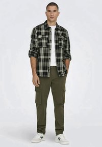 Camicia a quadri in bianco e nero, sovrapposta a una t-shirt bianca. Pantaloni cargo oliva con tasche laterali e sneakers bianche con dettagli verdi.