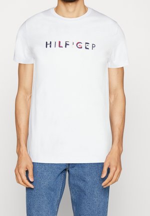 Mand iført hvid T-shirt med korte ærmer og flerfarvet "HILFIGER"-tekst samt blå jeans, stående mod en ensfarvet hvid baggrund.