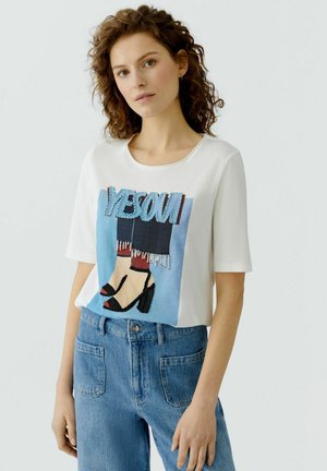 Frau mit lockigem Haar trägt ein weißes T-Shirt mit buntem Motiv von Beinen in schwarzen High Heels und blauen Jeans vor schlichtem Hintergrund.
