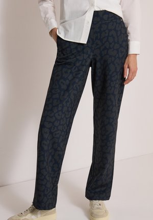 Pantalon bleu foncé avec un imprimé léopard subtil, porté avec une chemise blanche et des baskets beige, une main dans la poche, debout devant un fond clair.