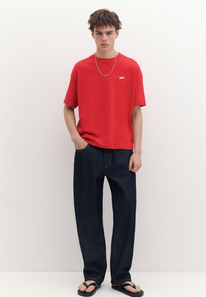Ein junger Mann steht an einer weißen Wand, trägt ein rotes T-Shirt, dunkelblaue Jeans, schwarze Flip-Flops und eine silberne Halskette.